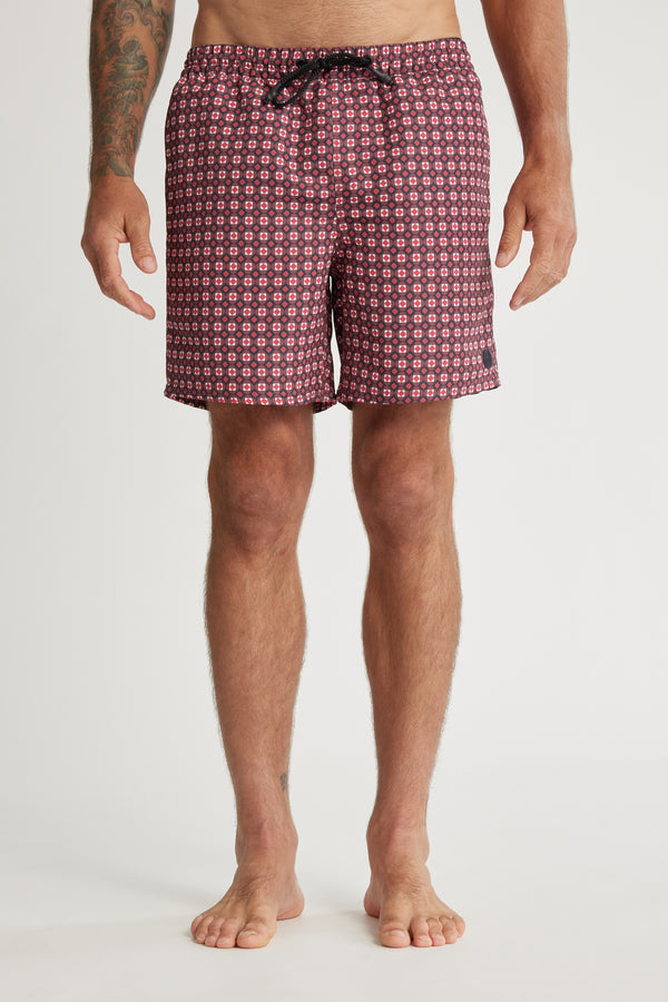 A person stands barefoot against a plain white background wearing Barkers Laramie Print Volley shorts—red and black patterned, recycled polyester with a drawstring waist. Their arms are relaxed by their sides, one displaying a tattoo.