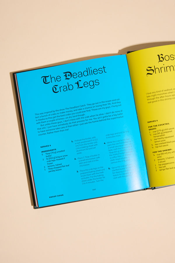 An open From Crook to Cook cookbook by Other rests on a beige surface, showing a blue page titled "The Deadliest Crab Legs" with Snoop Dogg's recipes and ingredients, and part of a yellow seafood recipe page on the right.