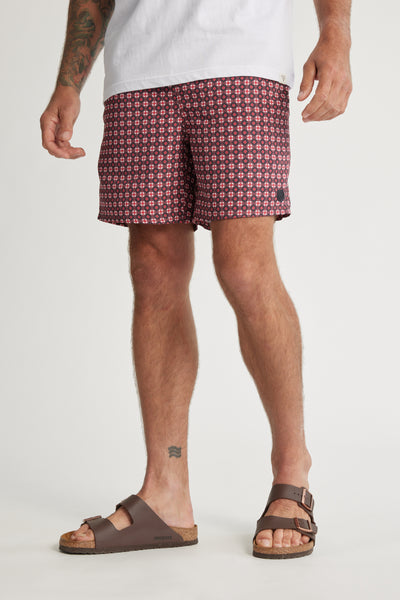 A man stands against a plain background wearing Barkers' Laramie Print Volley red geometric shorts, a white t-shirt, and brown sandals, displaying tattoos on his leg and arm.