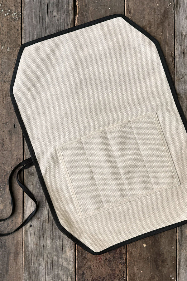 The IronClad Ghost Knife Roll, featuring beige fabric, black edging, and four stitched pockets, is displayed open on a rustic wooden surface.