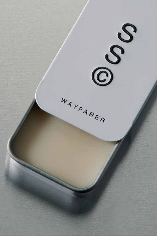 A rectangular metal tin labeled "Solid State - Wayfarer" with a partially open sliding white lid reveals the wax-based solid cologne balm inside, set on a smooth, light gray surface.