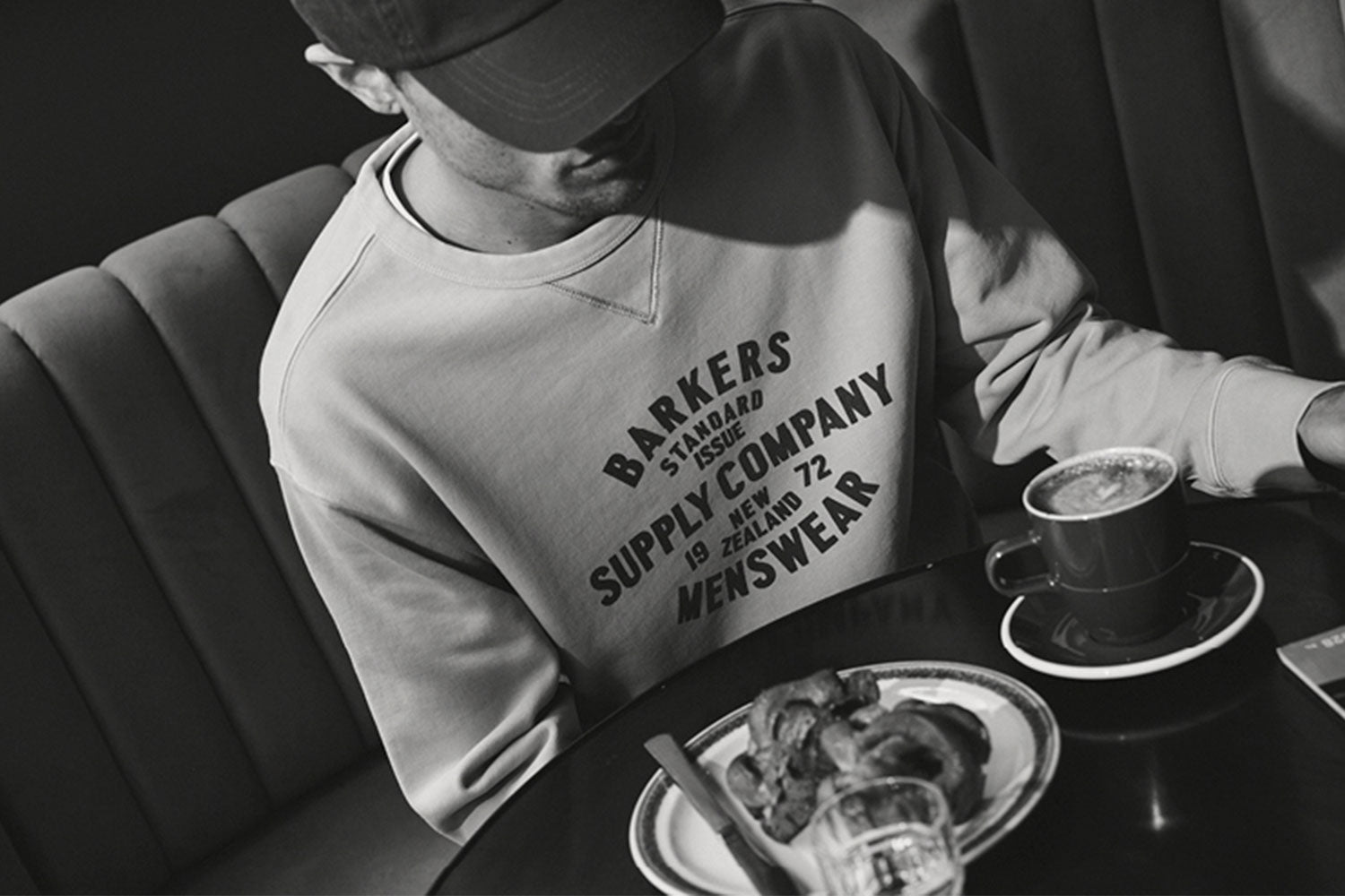 A man in a sweatshirt and cap sits at a café table with a coffee, a pastry on a plate, and a glass of water. The image is in black and white.