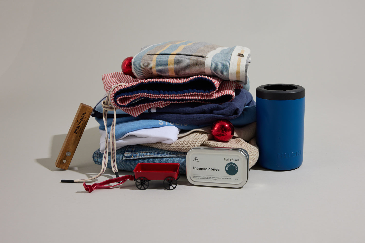 A neatly folded stack of casual clothes including jeans, shirts, and a red striped garment, topped with a beige baseball cap with a small embroidered logo, all set against a neutral background.