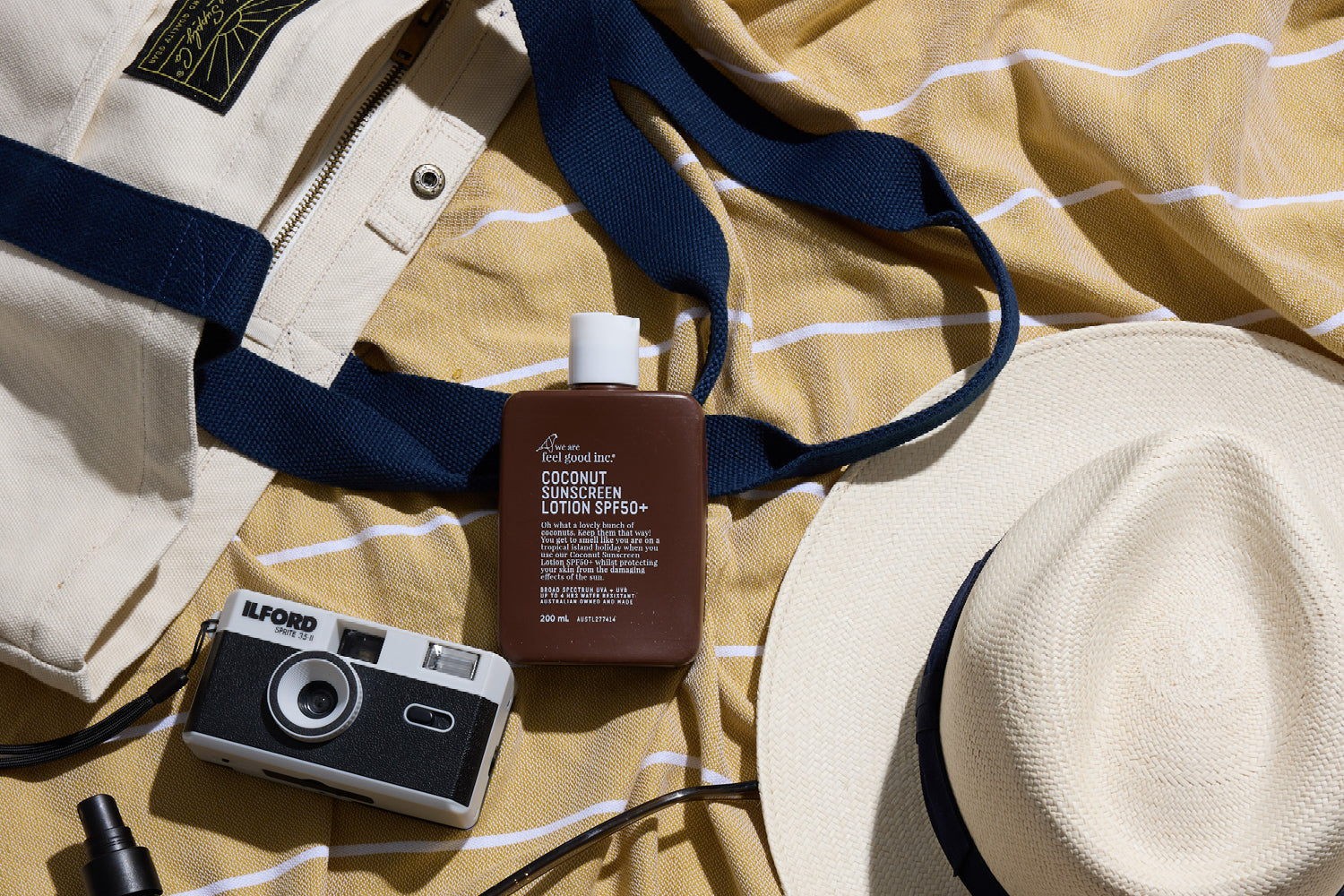 A flat lay of summer essentials on a yellow striped towel: a white tote bag with blue straps, a brown bottle of sunscreen, a white sun hat, and a vintage Ilford film camera.
