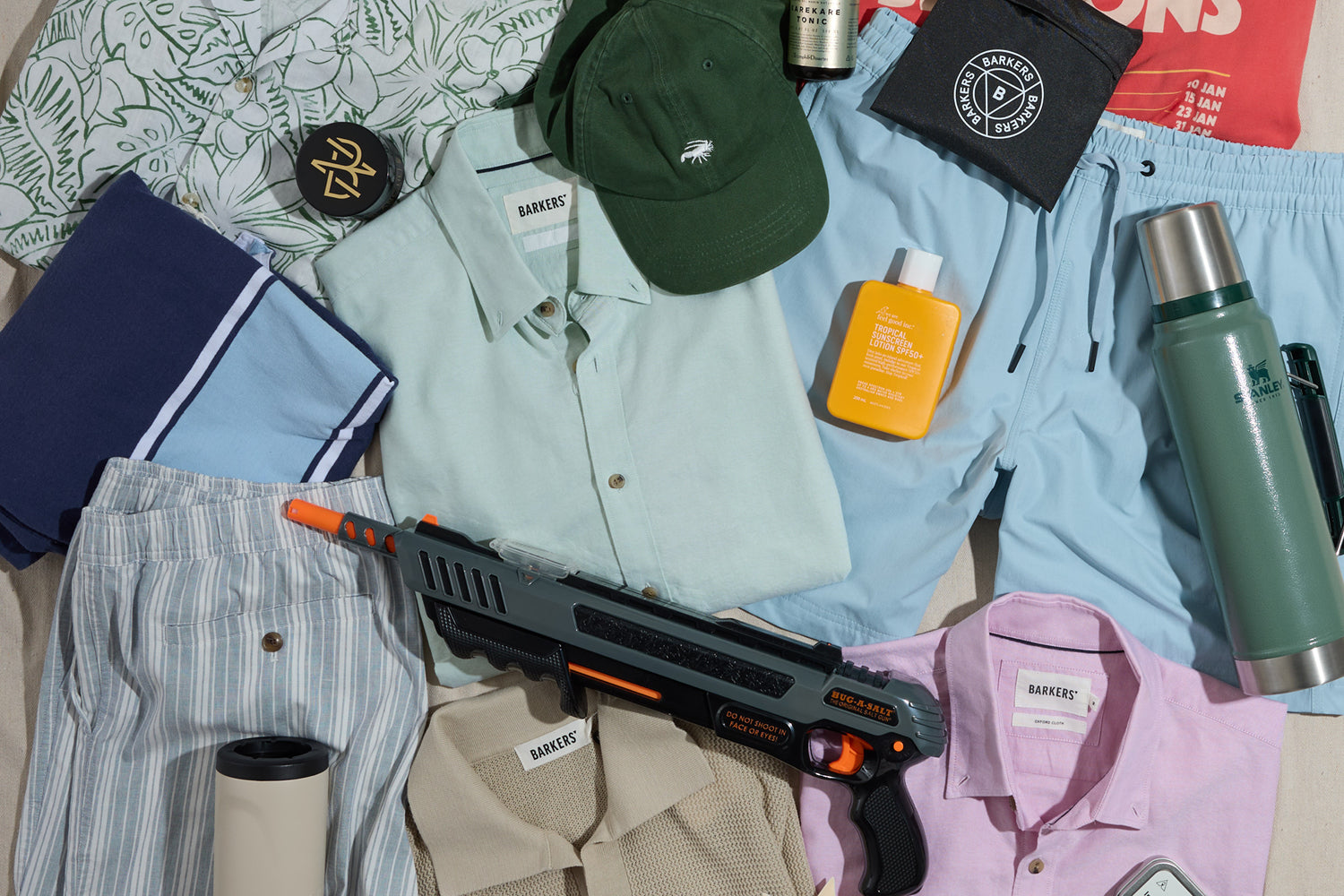 A flat lay of summer clothing, a hat, water bottles, sunscreen, a thermos, a pool towel, and a toy gun arranged together on a light surface.