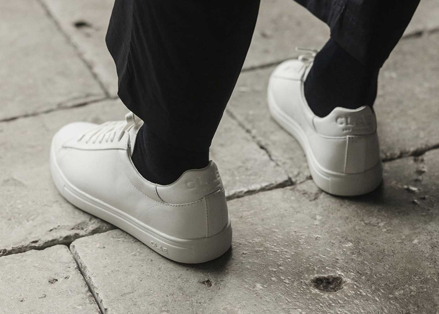 Person wearing white sneakers with black socks and black pants, standing on a large stone tile floor. The sneakers have a minimal, clean design.