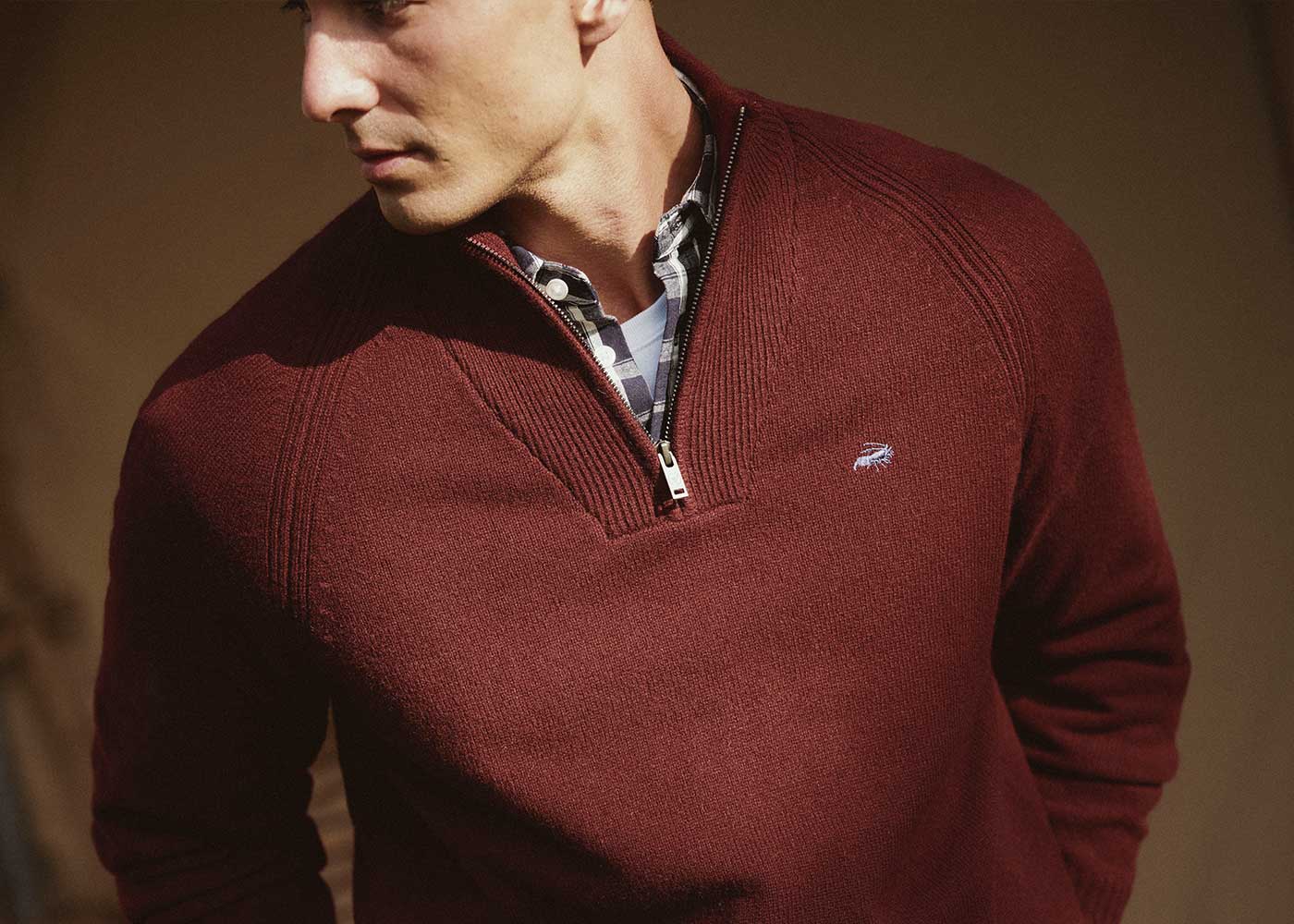 A man wearing a maroon quarter-zip sweater over a plaid shirt looks to the side. The sweater features a small embroidered logo on the chest.