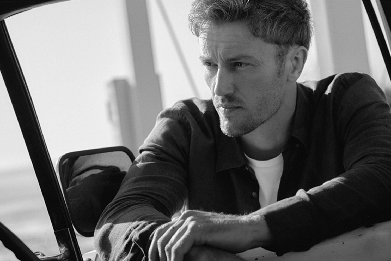 A man with short, wavy hair leans on the open window of a vehicle, gazing thoughtfully into the distance in a black and white photo.