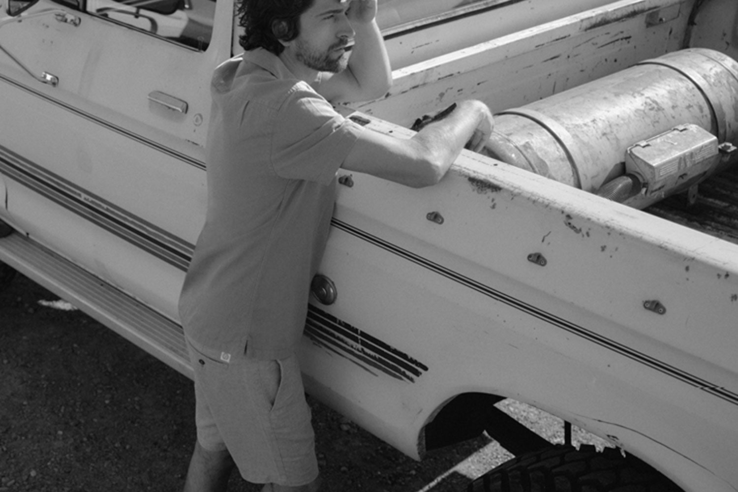 A man in casual clothes leans on the side of an old pickup truck, looking thoughtfully into the distance. The photo is in black and white.