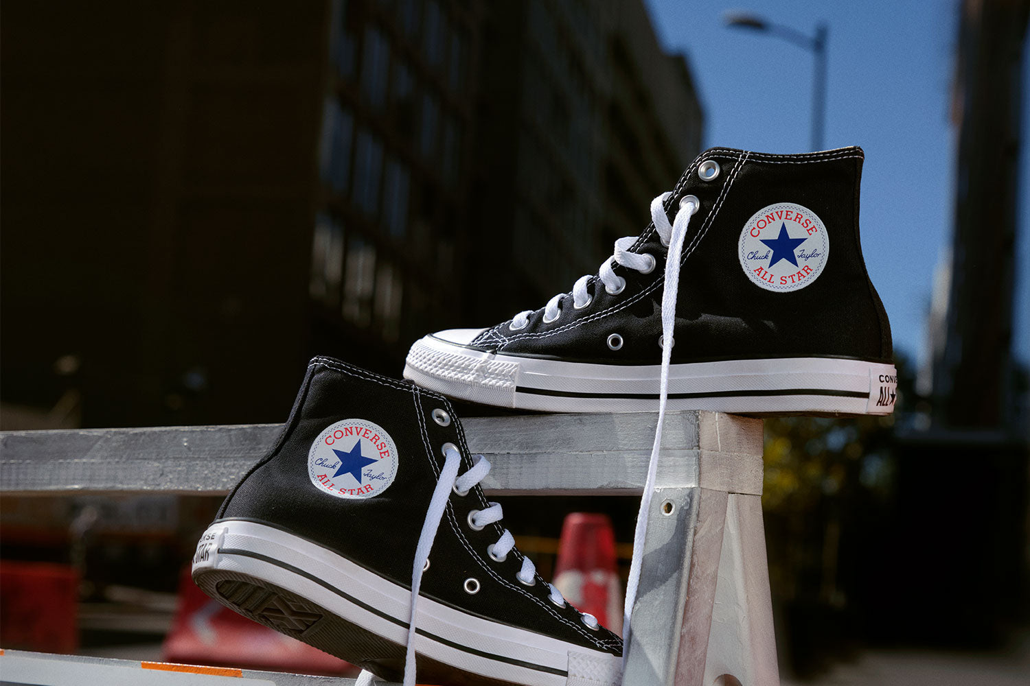 A pair of black Converse All Star high-top sneakers rest on a metal rail outdoors, with city buildings and a blue sky blurred in the background.