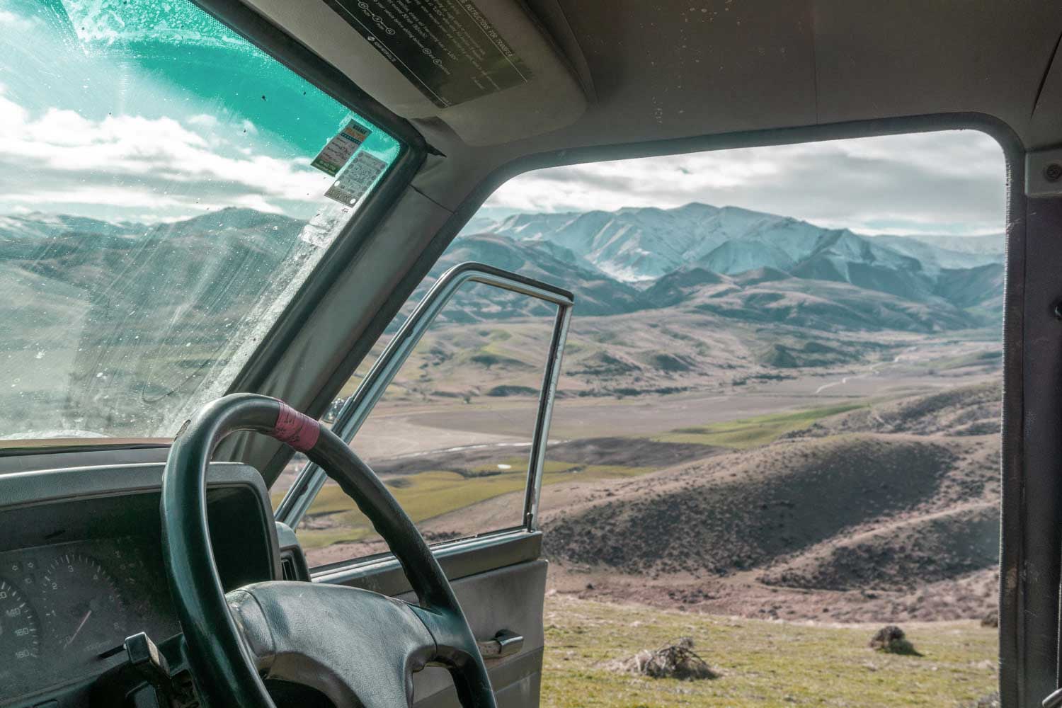 View from inside a vehicle with the driver’s door open, looking out onto a scenic landscape of rolling hills and distant snow-capped mountains under a partly cloudy sky.