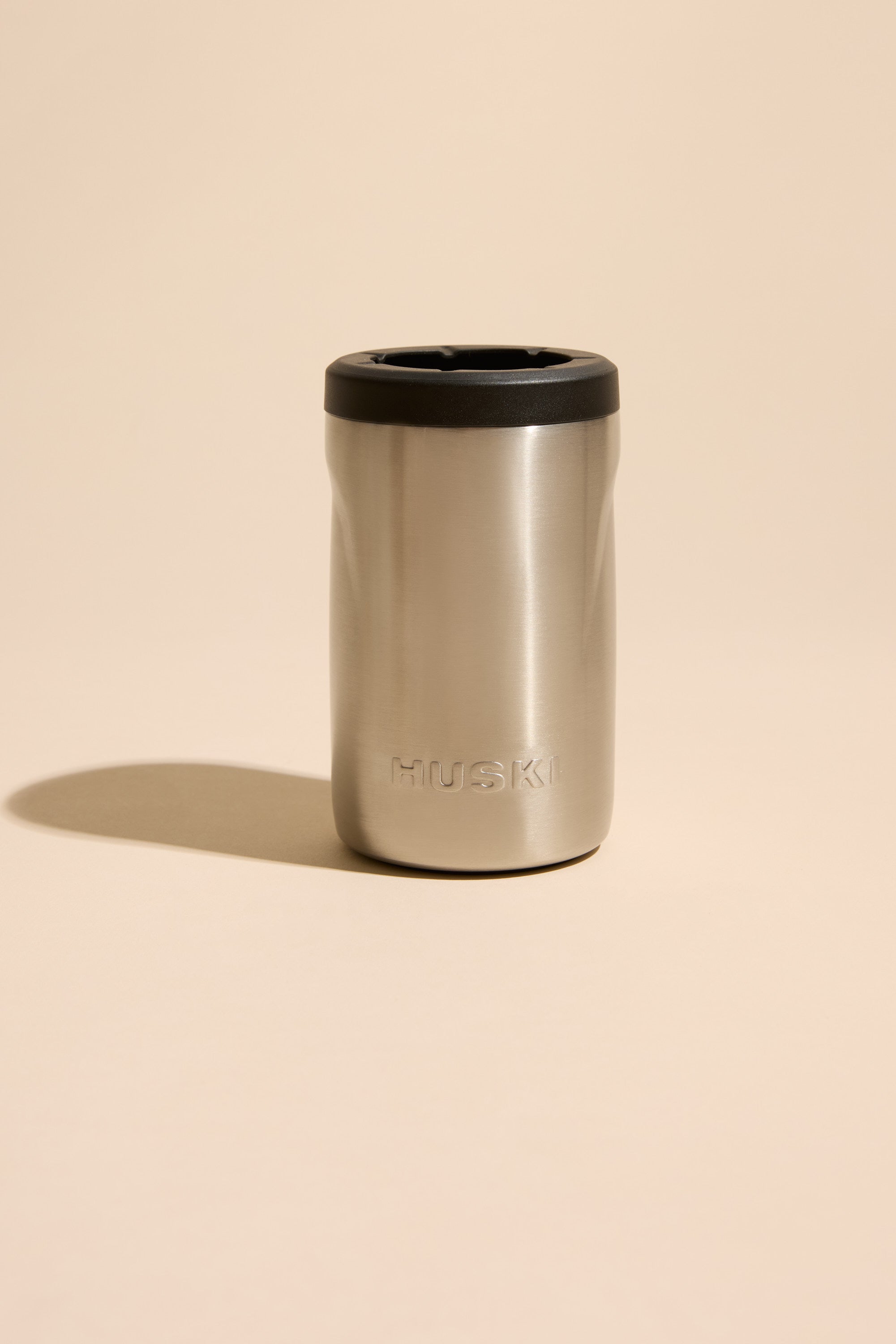 The Huski Beer Cooler by Huski is a high-performance stainless steel beer cooler with a black lid, embossed branding on the front, shown standing upright on a beige background with a soft shadow to the left.