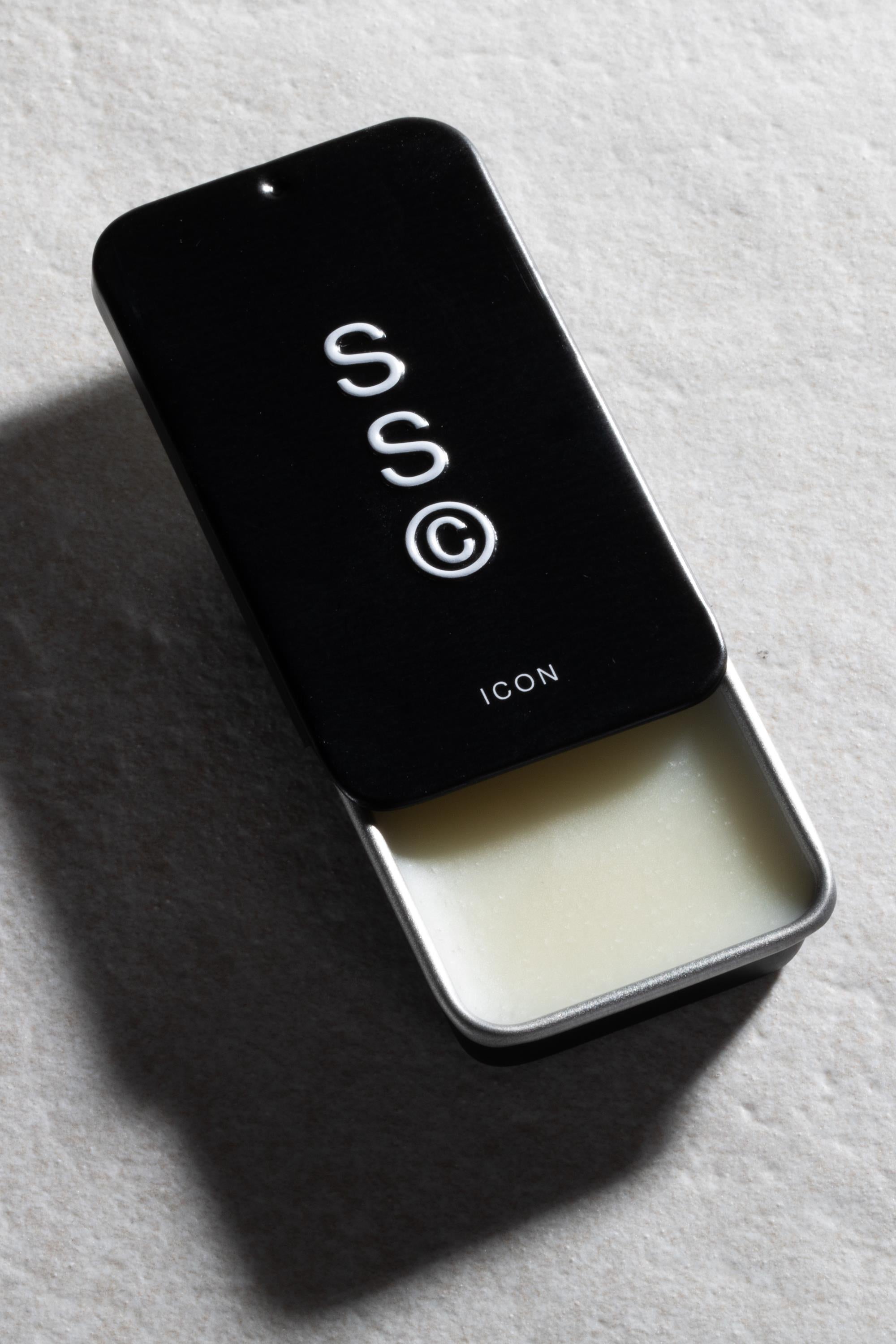 A rectangular black Solid State - Icon tin, its lid partly open to reveal solid white wax-based cologne, rests on a light textured surface.