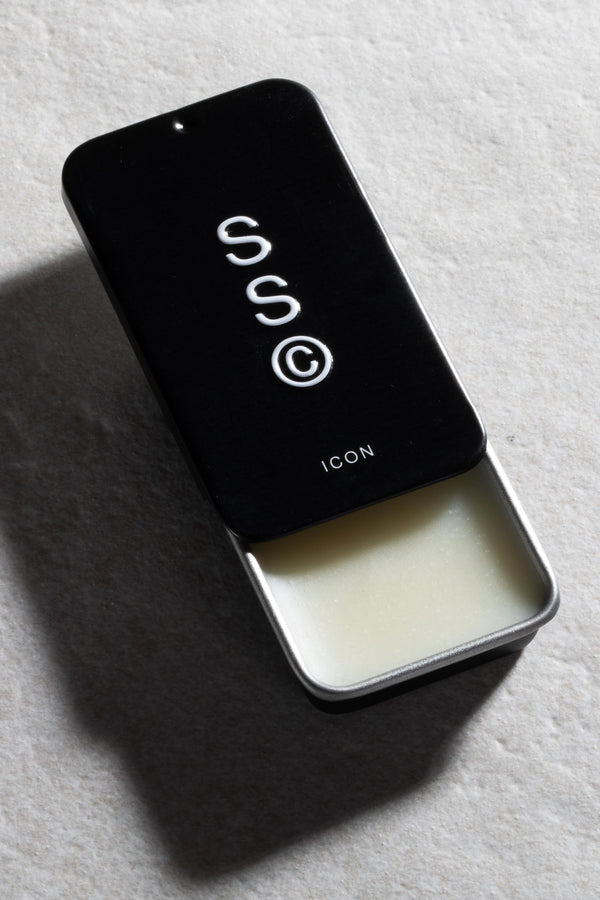 A rectangular black Solid State - Icon tin, its lid partly open to reveal solid white wax-based cologne, rests on a light textured surface.