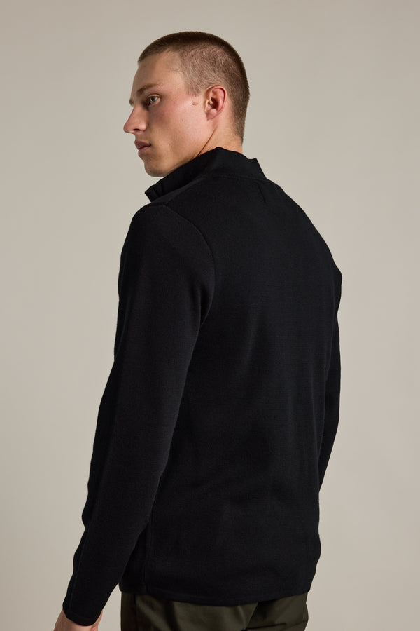 A young man with short hair stands facing away, looking over his left shoulder, wearing the Barkers Brandon ZQRX Merino 1/4 Zip—an ideal black layering piece for cold weather—against a plain light background.