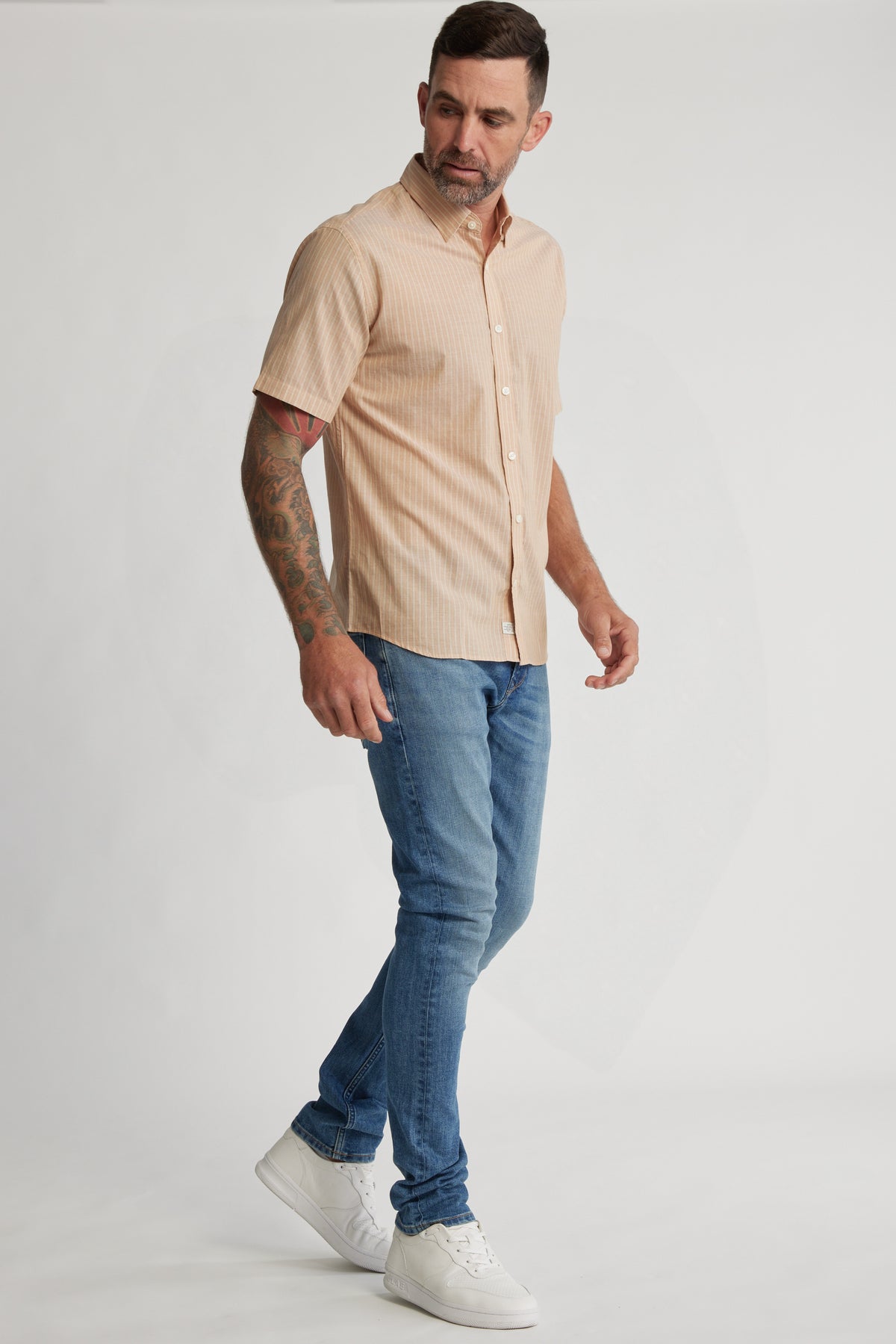 A man in a tailored fit Barkers Burnham Stripe Shirt, blue jeans, and white sneakers stands against a plain white background, looking down to his left with one arm slightly raised.