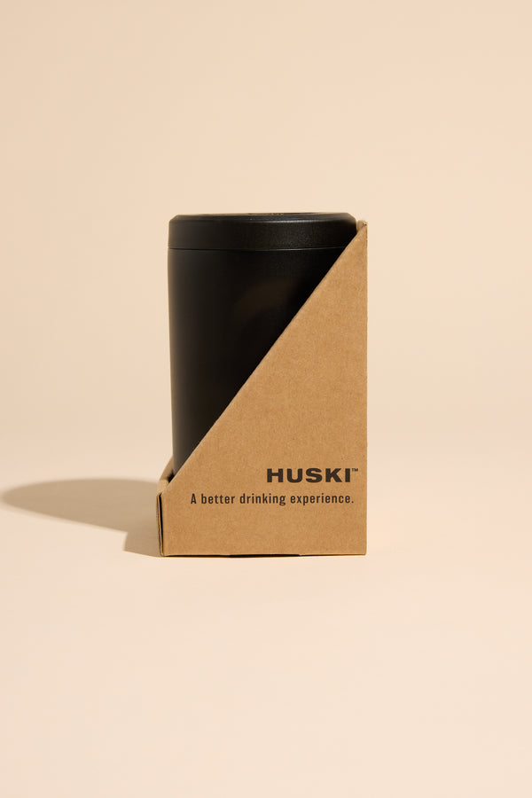 A black stainless steel Huski Beer Cooler by Huski is partially enclosed in a minimalist brown cardboard sleeve with “HUSKI – A better drinking experience” printed on it, set against a plain beige background to highlight its high-performance cooling.