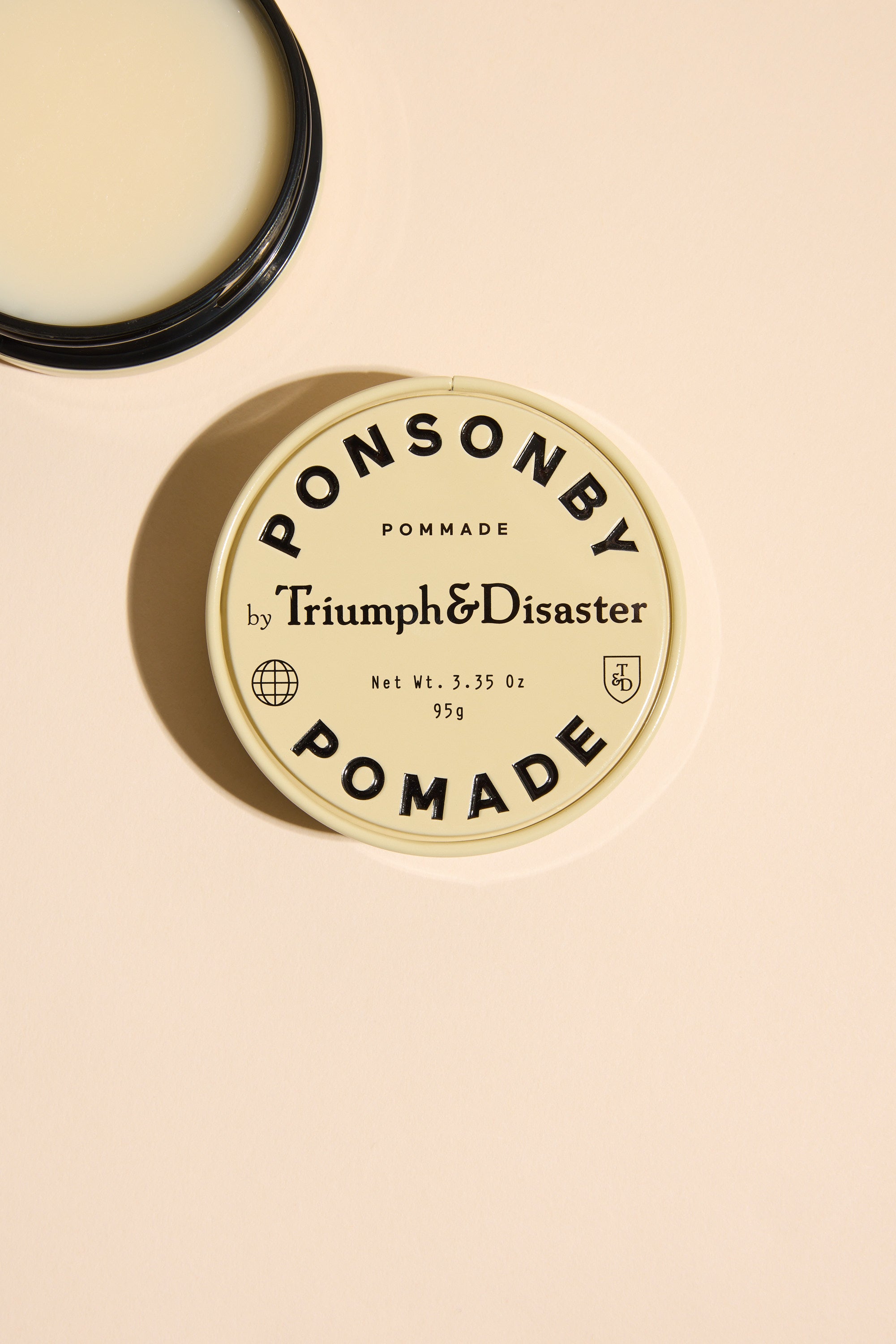 A tin of Triumph & Disaster Ponsonby Pomade, a natural hair pomade by Triumph & Disaster, sits on a light beige surface with its lid off, revealing the product and its bold black label with a minimalist design.