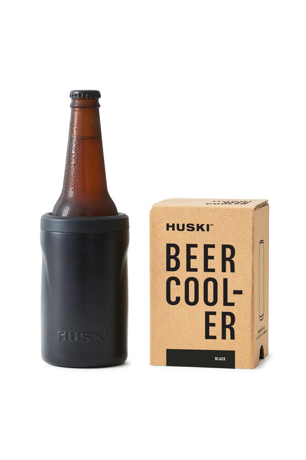 A brown beer bottle in a black Huski Beer Cooler by Huski sits beside a branded cardboard box, all on a white background.