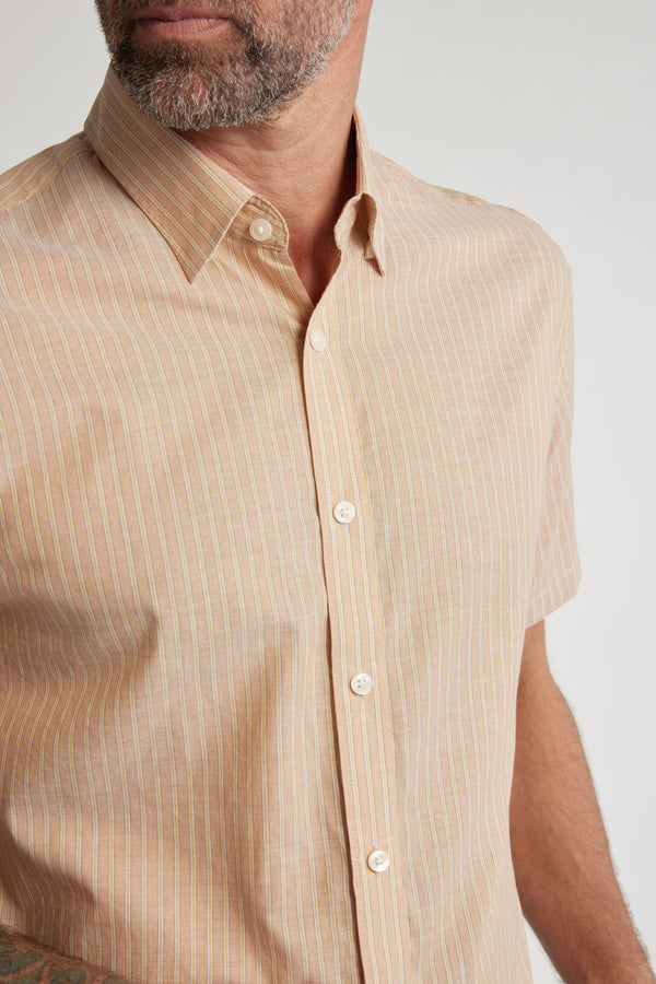 A man with a short beard wears the Barkers Burnham Stripe Shirt, a light brown, short-sleeve, button-up made of lightweight cotton with thin white stripes and a tailored fit, standing against a plain background.