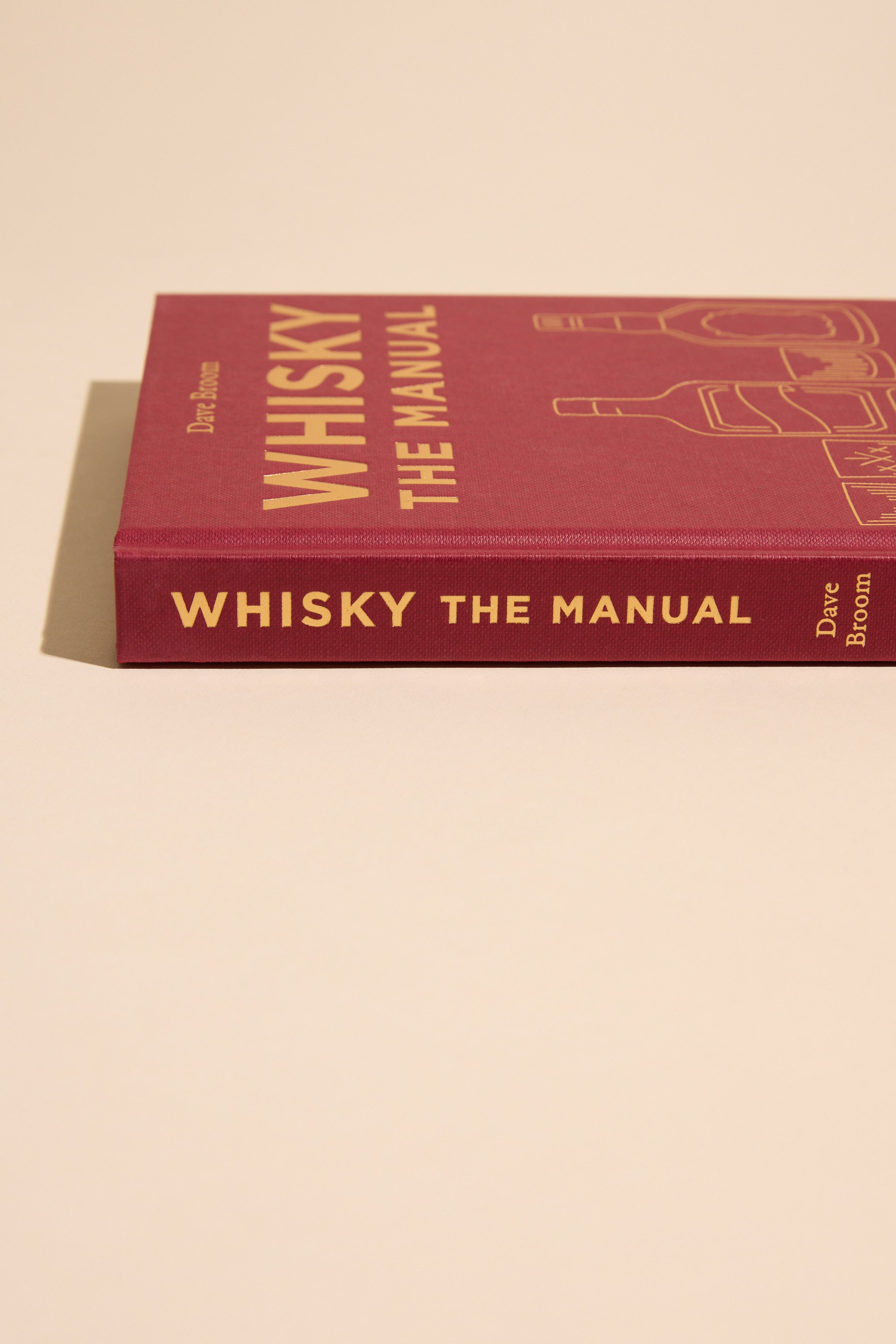 A maroon hardcover copy of Whisky The Manual by Other rests on a beige surface, displaying gold lettering and whisky bottle outlines—an essential guide featuring expert whisky tasting notes.