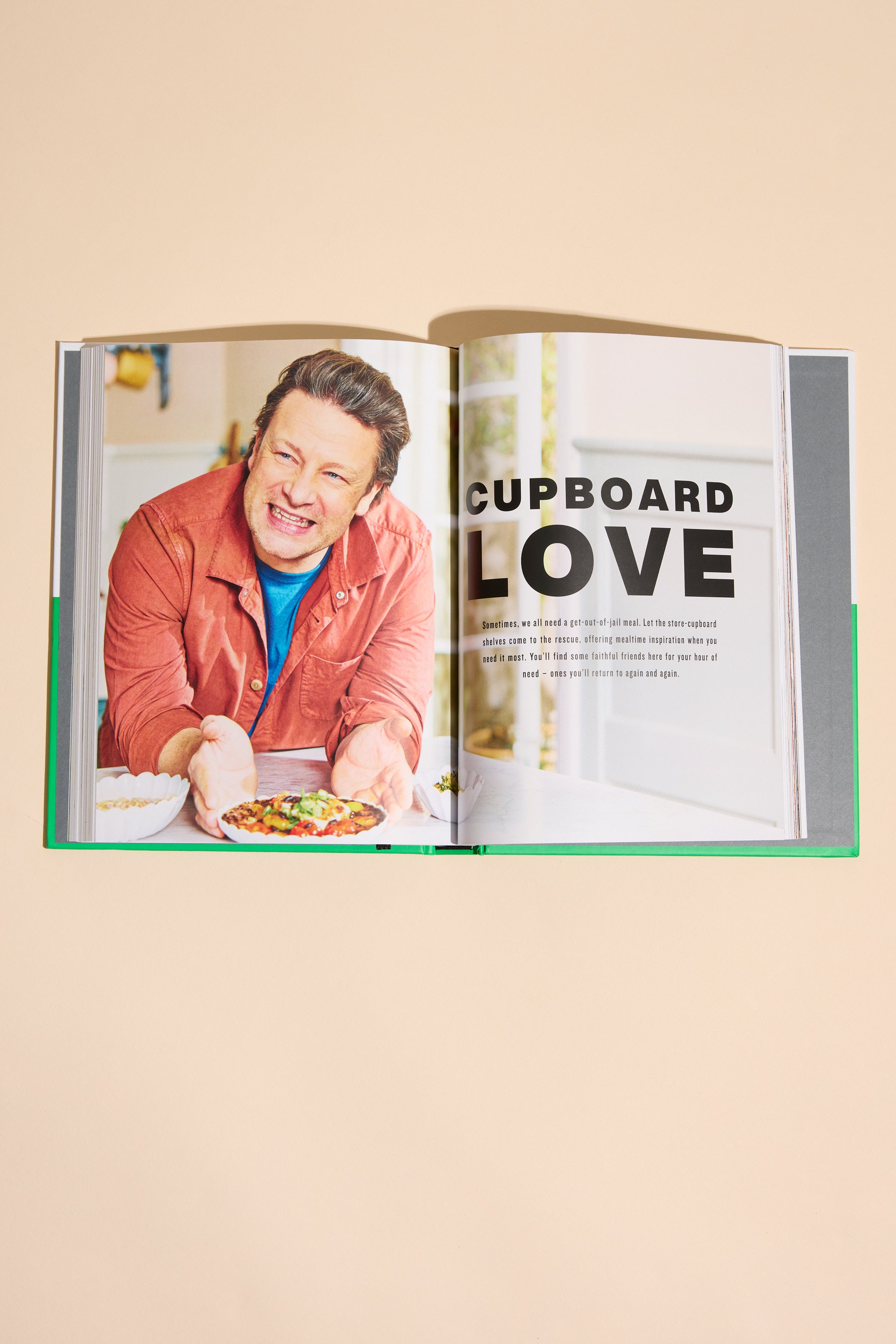 An open Simply Jamie cookbook from Other shows a smiling man in an orange shirt holding a bowl of food, with “CUPBOARD LOVE” as the heading on the right page above a paragraph about easy recipes.