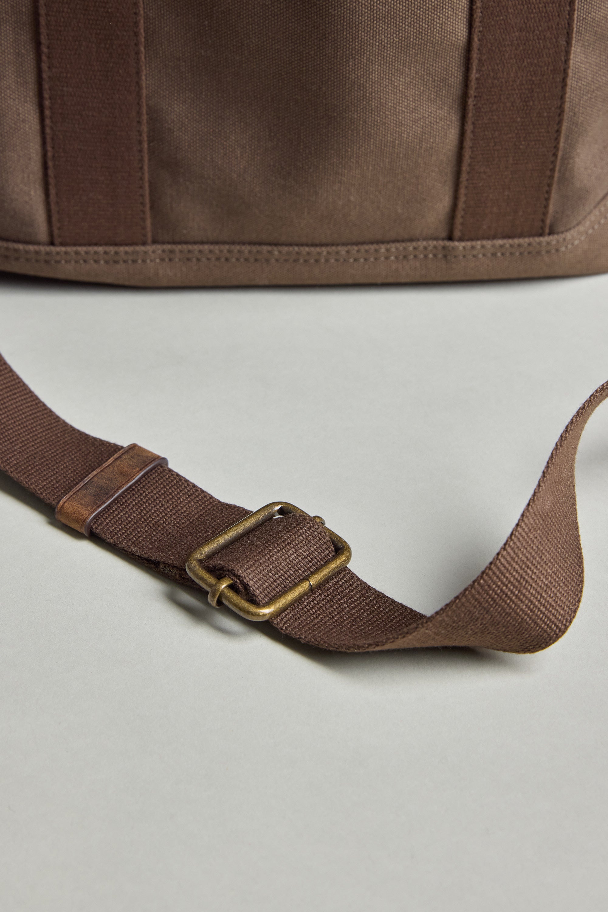 The Barkers Foxton Canvas Convertible Tote is shown in close-up—brown cotton canvas with a matching adjustable strap and brass-tone buckle—laid on a neutral surface. Versatile, it seamlessly transitions from tote to backpack for everyday style.