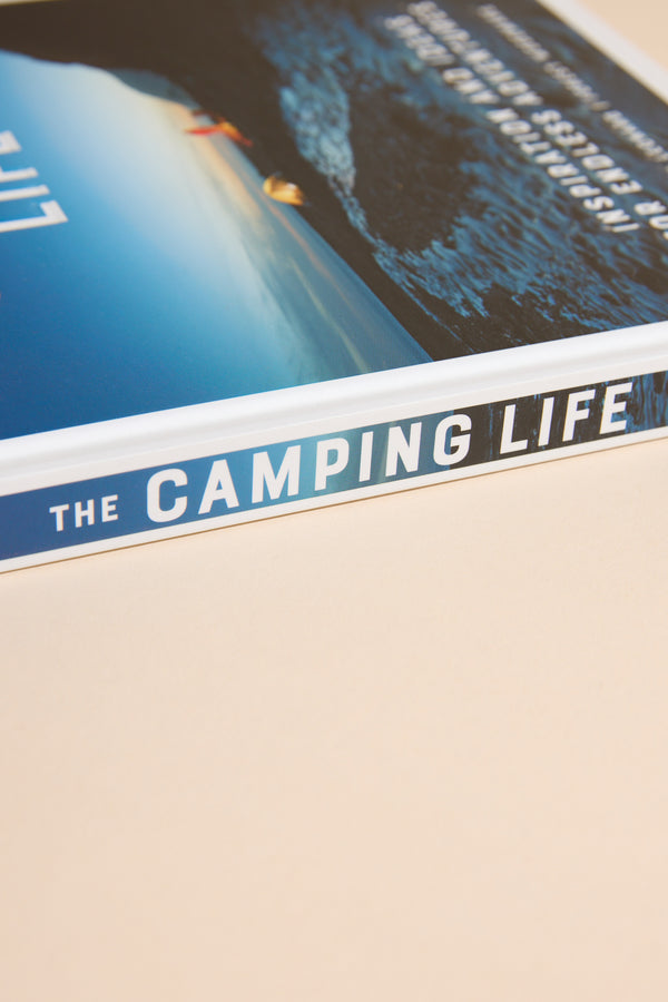 A close-up of the spine of Camping Life by Other on a light beige surface, featuring a scenic outdoor adventure cover with tents pitched beside a tranquil New Zealand lake.