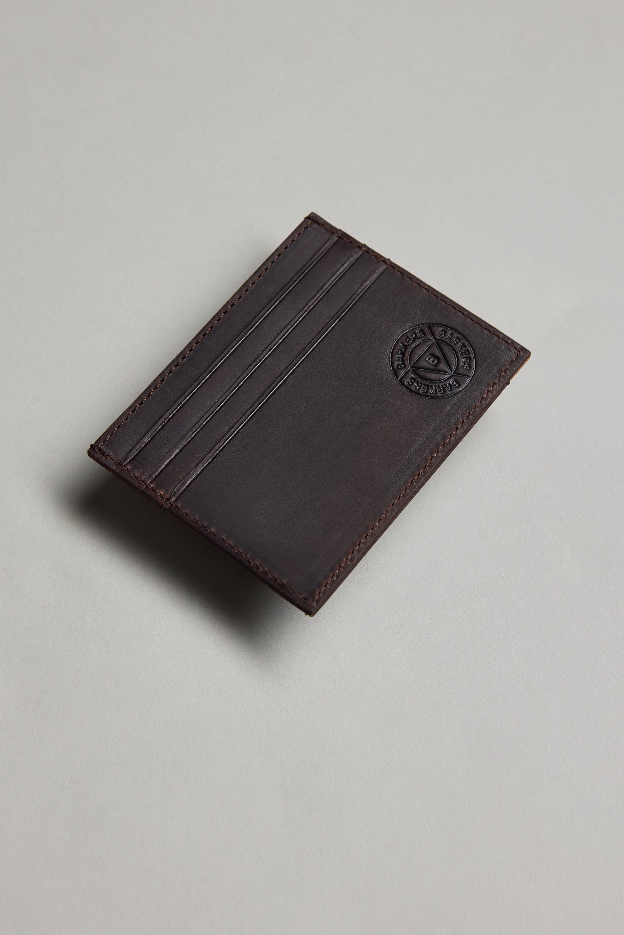 The Barkers Taylor Leather Card Wallet, in dark brown slim leather with three card slots and an embossed circular logo, made from Leather Working Group-certified materials, is shown on a light grey surface.