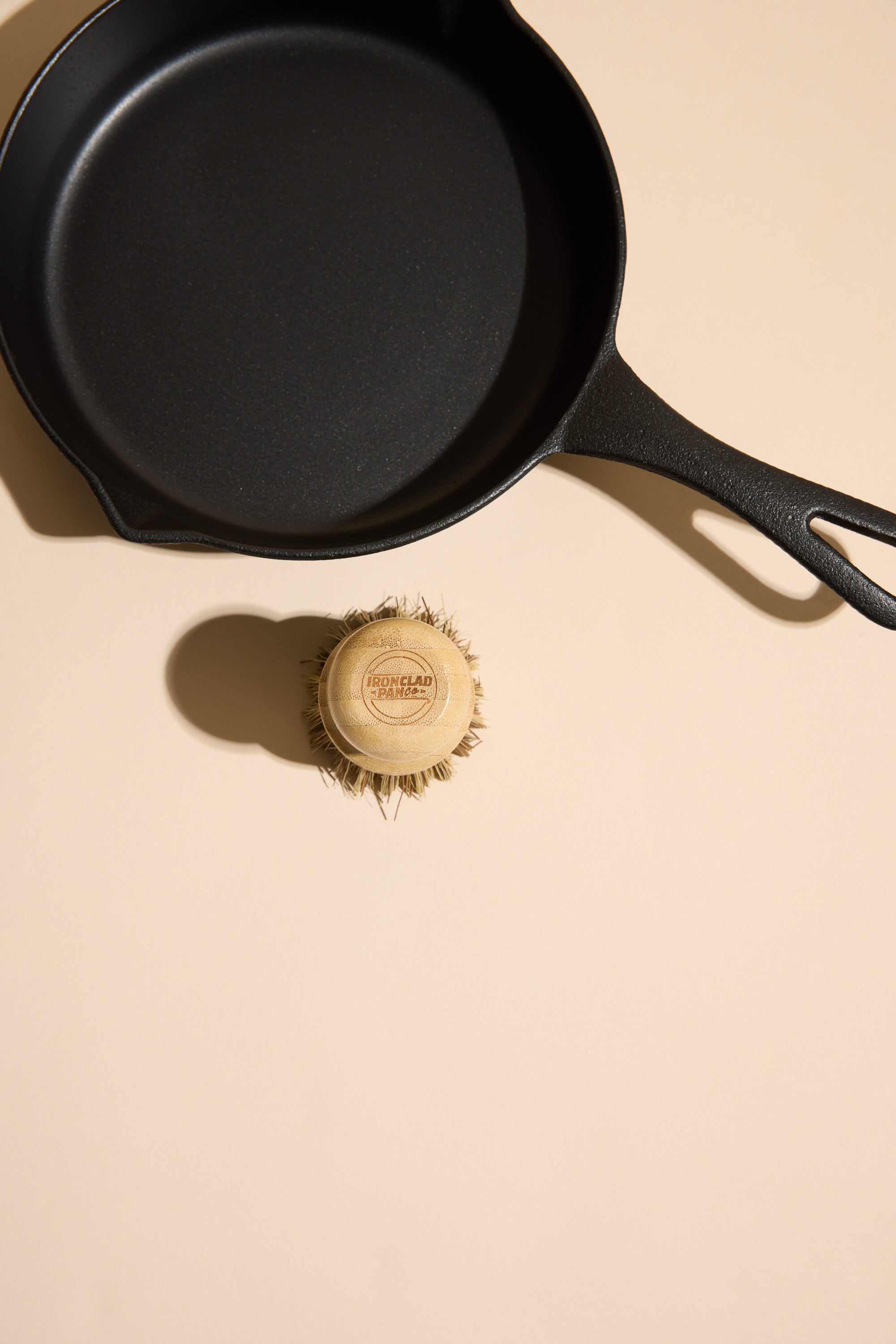 The Ironclad Lil' Legacy Pan by Other sits on a light beige surface next to a round bristle scrub brush with a wooden handle labeled IRONCLAD CAST IRON CARE.
