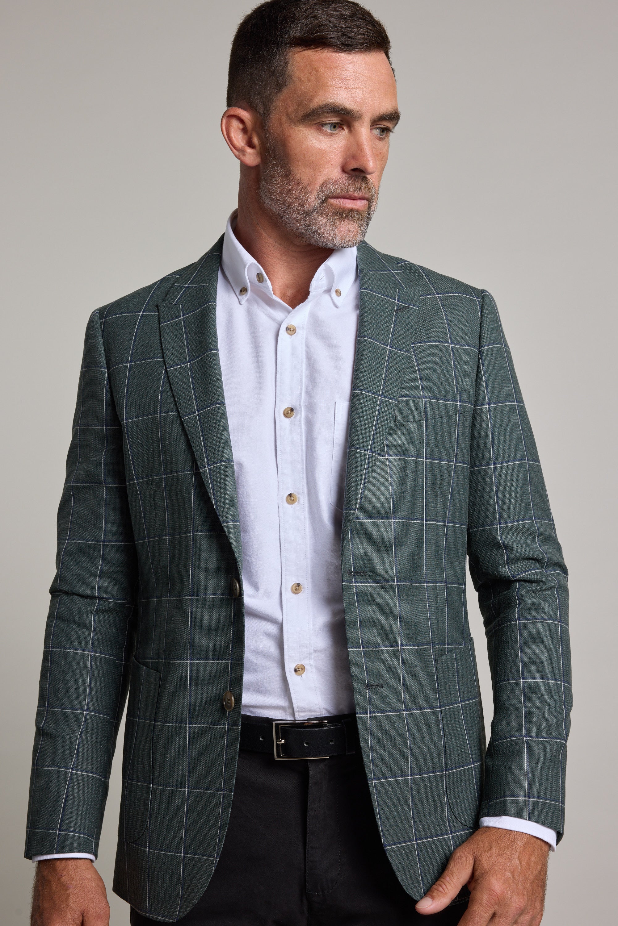 A man with short brown hair and a beard models the Barkers Nathaniel Check Blazer in green over a white shirt and black pants, standing against a plain light background and gazing to his left.