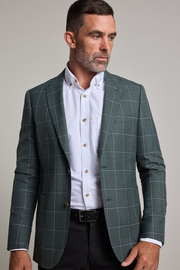 A man with short brown hair and a beard models the Barkers Nathaniel Check Blazer in green over a white shirt and black pants, standing against a plain light background and gazing to his left.