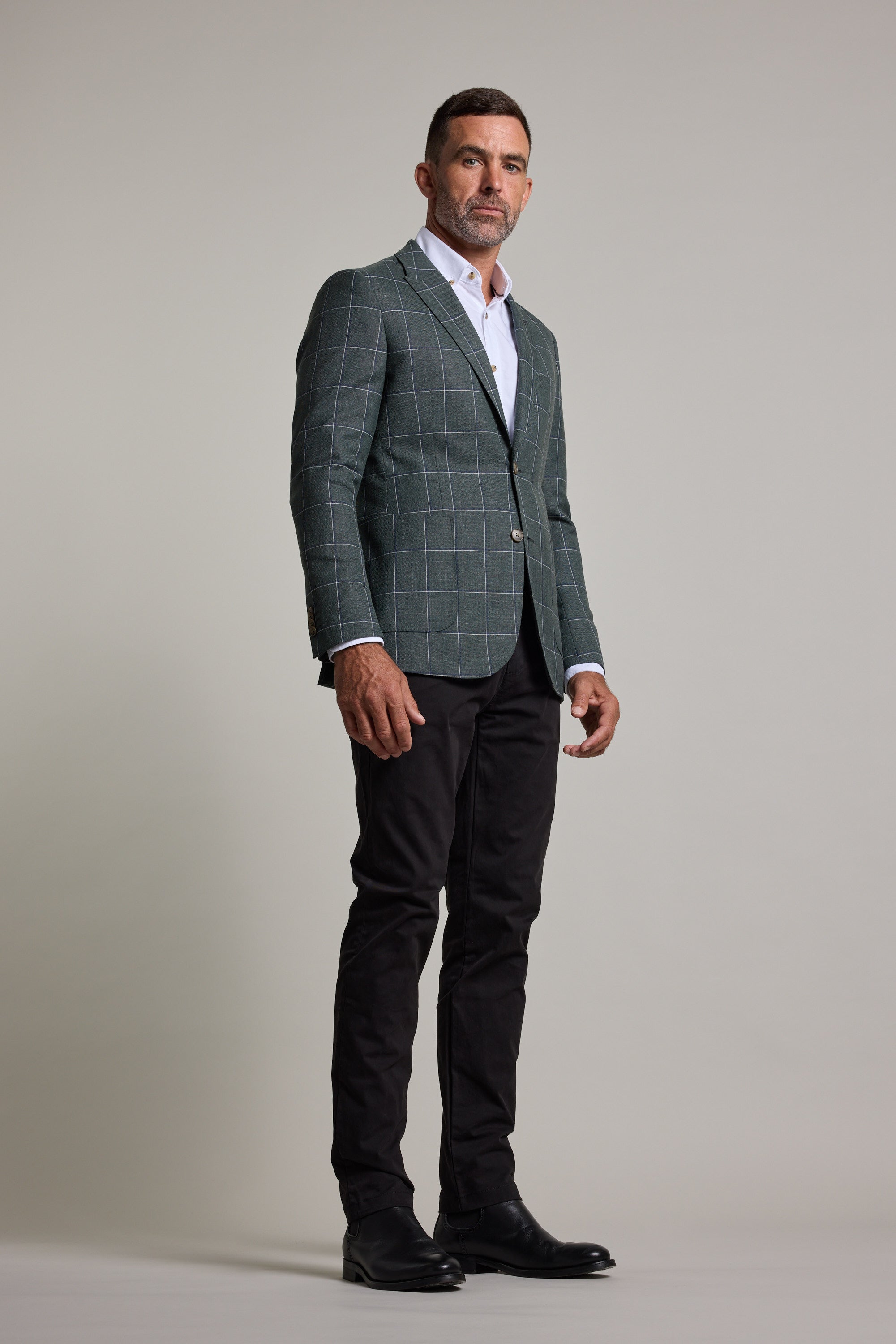 A man with short dark hair and a beard stands confidently in the Barkers Nathaniel Check Blazer—a green wool blend check blazer—paired with a white shirt, black pants, and black shoes against a plain light gray background.