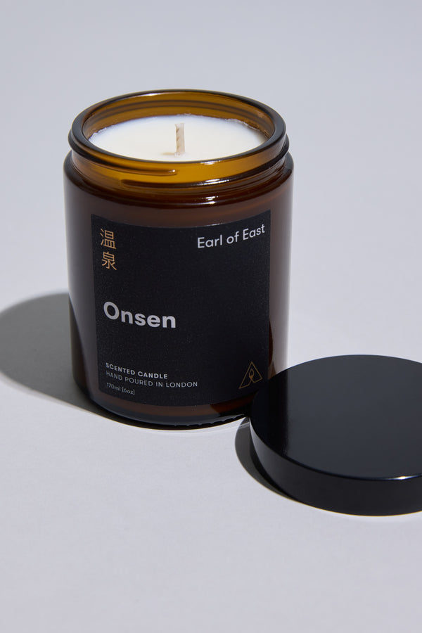 A brown glass jar EoE Onsen Candle 170ml by Earl of East, with a black lid beside it. The label shows English and Japanese text, and the white peppermint-scented soy wax is visible inside.