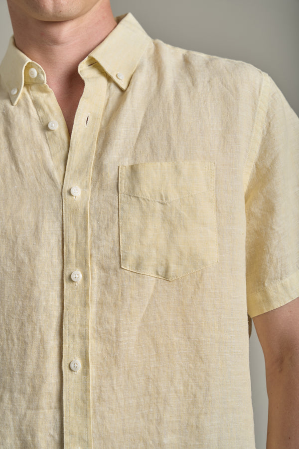 A close-up shows a person in the Barkers Gentry Linen Shirt—a light yellow, short-sleeved French linen shirt with button-down collar and chest pocket—perfectly lightweight and breathable for summer. The person’s head is partially out of frame.