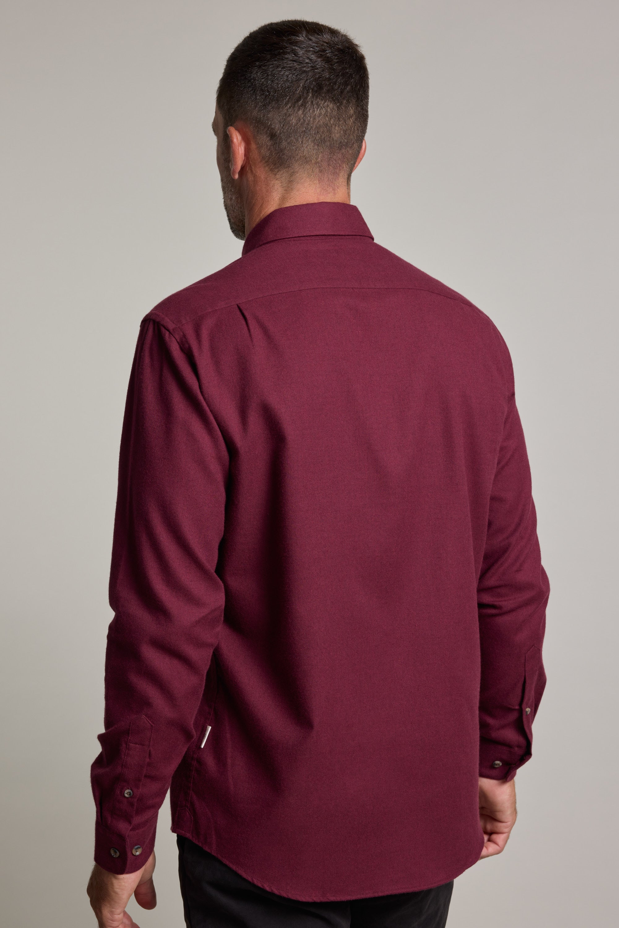 A man with short dark hair is seen from the back wearing a Barkers Antonio Shirt in maroon cotton-wool blend and black pants against a plain light gray background.