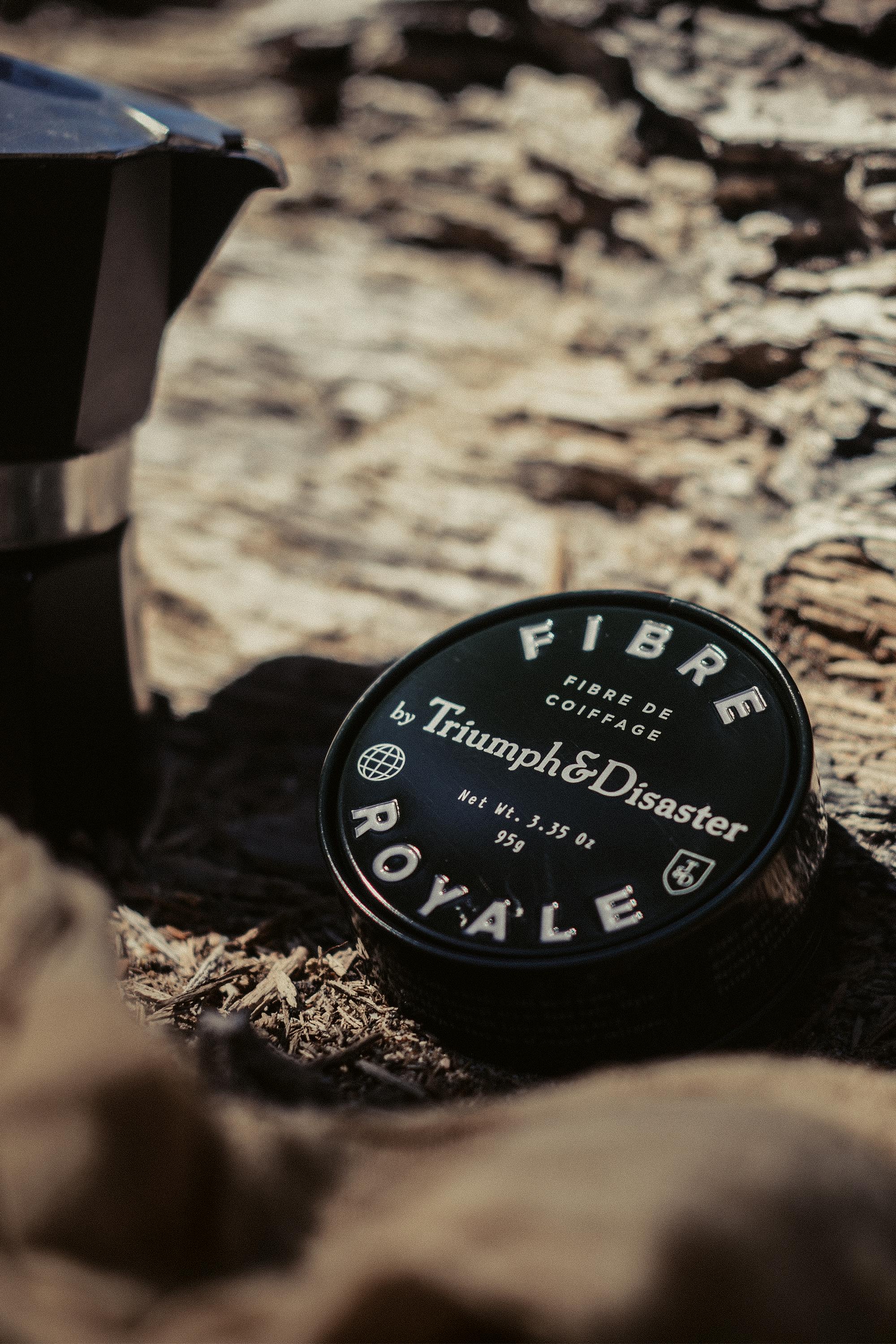 A black tin of Triumph & Disaster Fibre Royale sits on rough outdoor wood, partially framed by a metallic object on the left and a blurred beige cloth in front, highlighting this strong hold hair product.