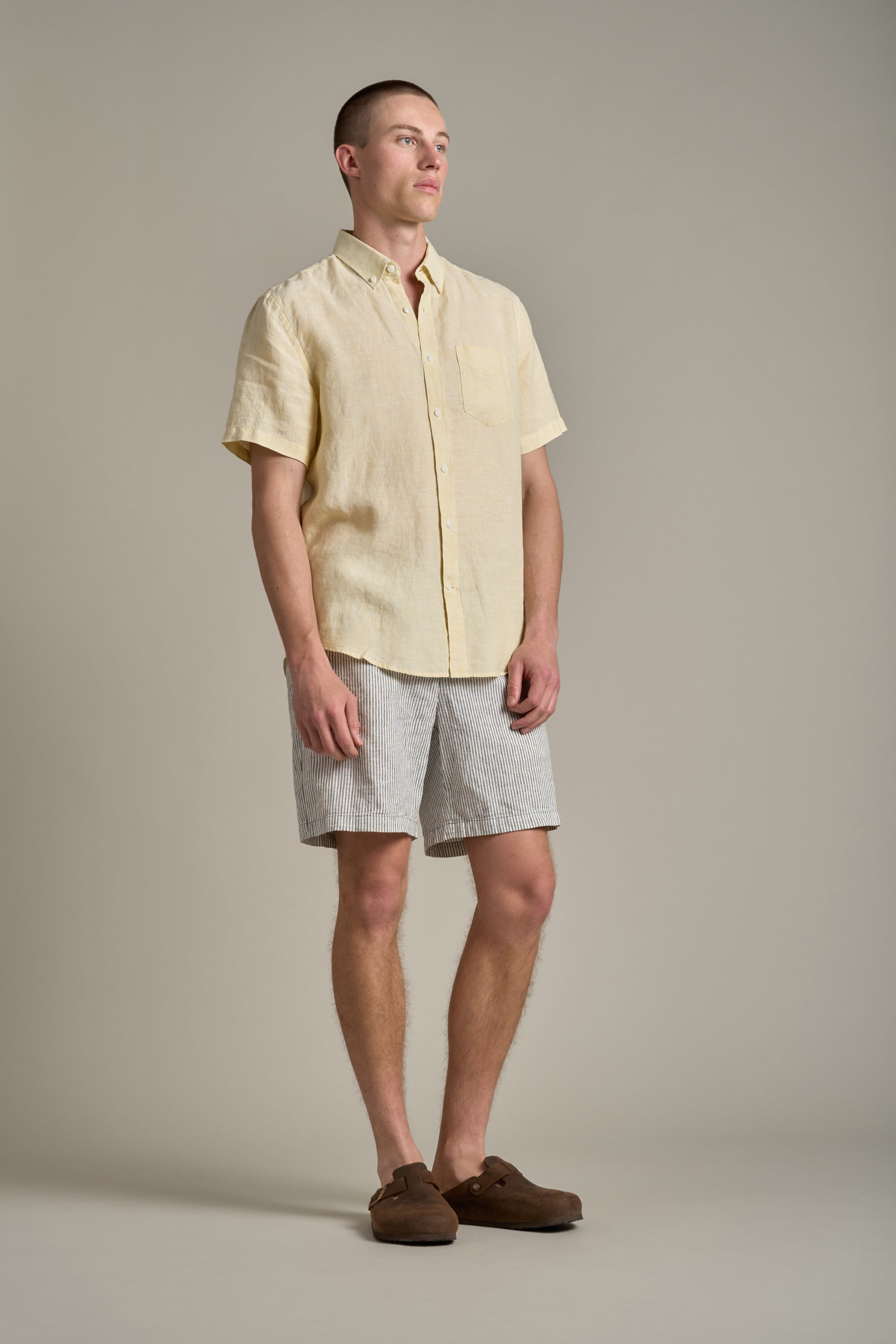 A man stands against a plain background wearing the Barkers Gentry Linen Shirt in yellow, paired with light gray striped shorts and brown slip-on shoes. He looks off to the side with a relaxed stance—ideal summer style.