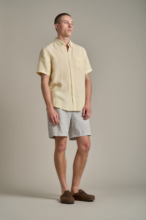 A man stands against a plain background wearing the Barkers Gentry Linen Shirt in yellow, paired with light gray striped shorts and brown slip-on shoes. He looks off to the side with a relaxed stance—ideal summer style.
