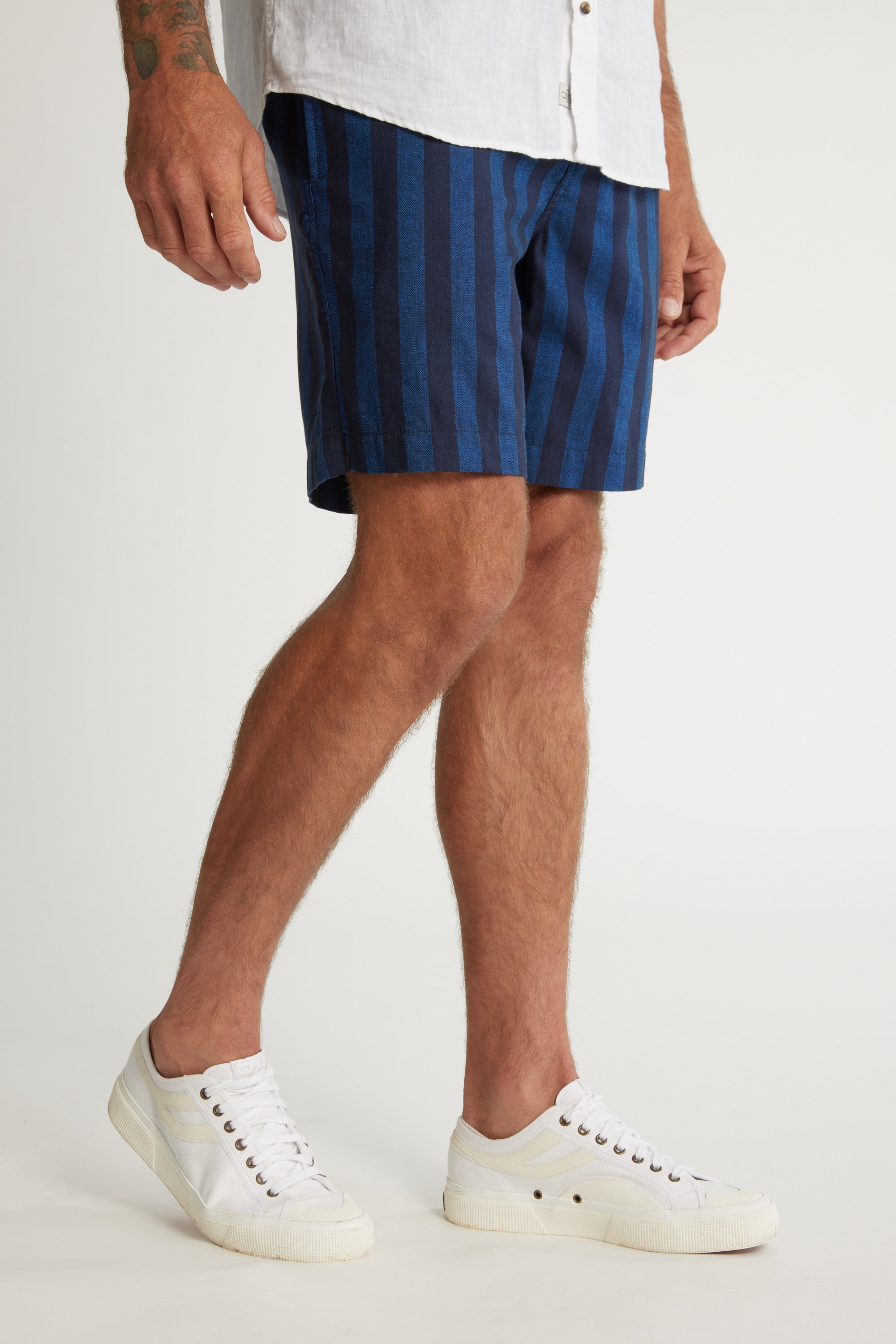 A man models Barkers Ashbury Stripe Short in blue and black with a white shirt and white sneakers against a plain background, showing only his lower body and one hand.