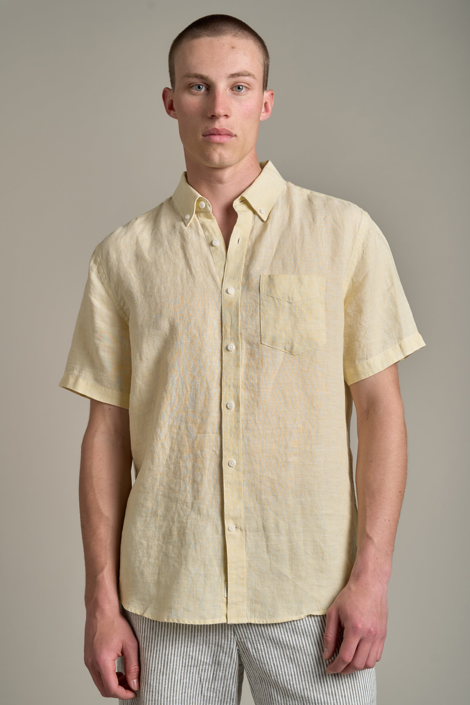 A young man with short hair models the Barkers Gentry Linen Shirt—a light yellow, breathable short-sleeve shirt with a chest pocket—paired with light striped pants, perfect for summer style against a plain background.