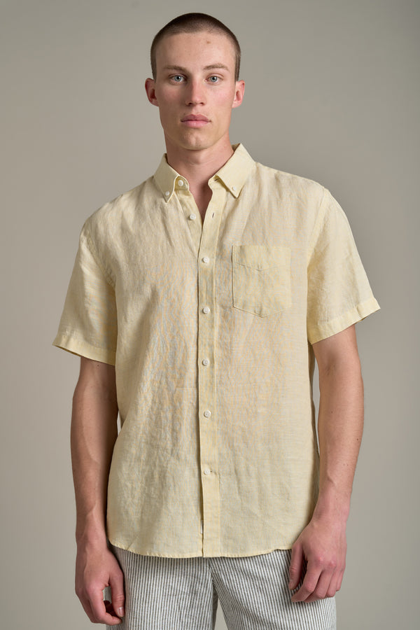 A young man with short hair models the Barkers Gentry Linen Shirt—a light yellow, breathable short-sleeve shirt with a chest pocket—paired with light striped pants, perfect for summer style against a plain background.