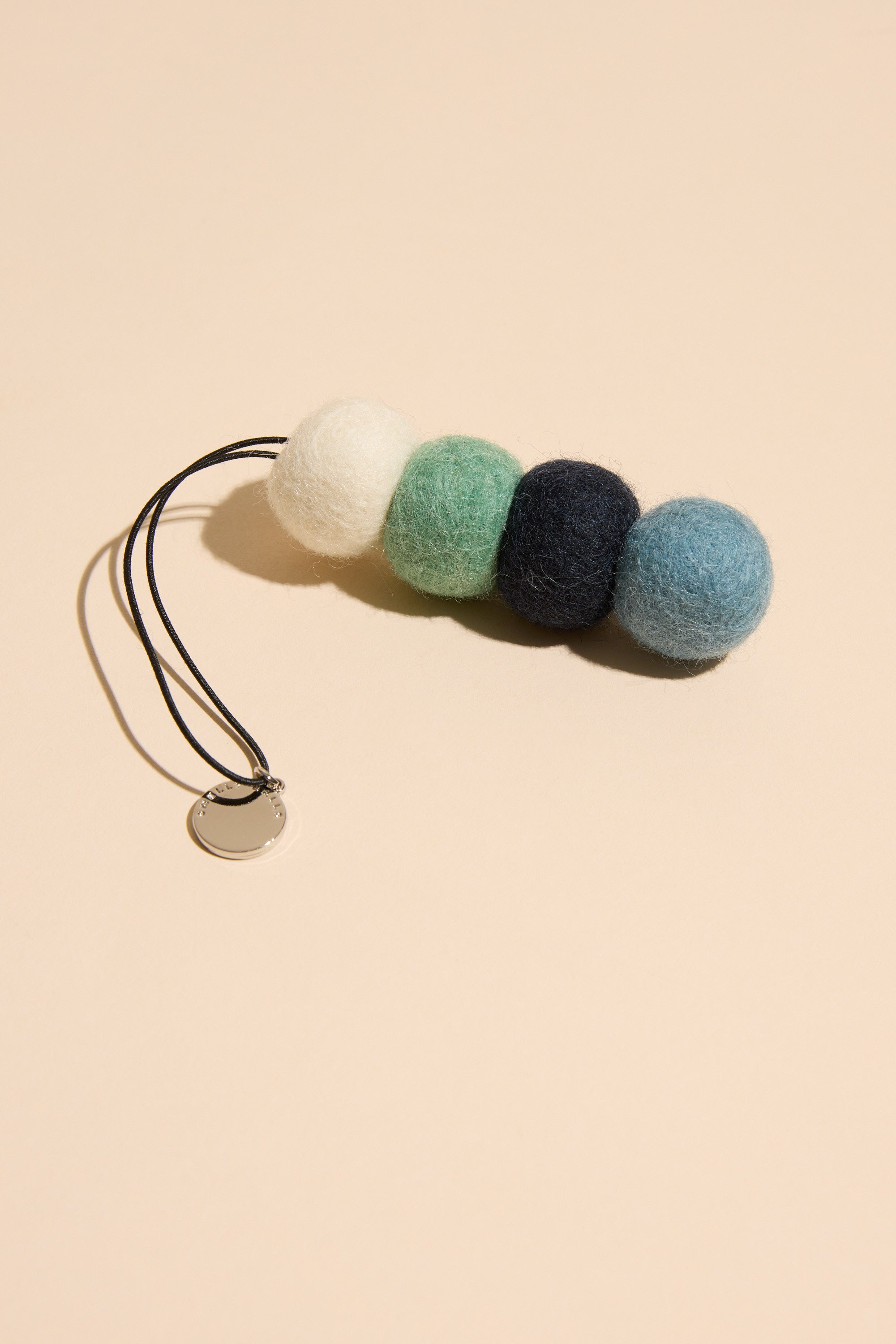 Smelly Balls: Cove / Coastal Drift by Other is a reusable car air freshener keychain with four felted wool balls in white, green, dark blue, and light blue, plus a round metal tag on a black cord. Photographed on a beige background.