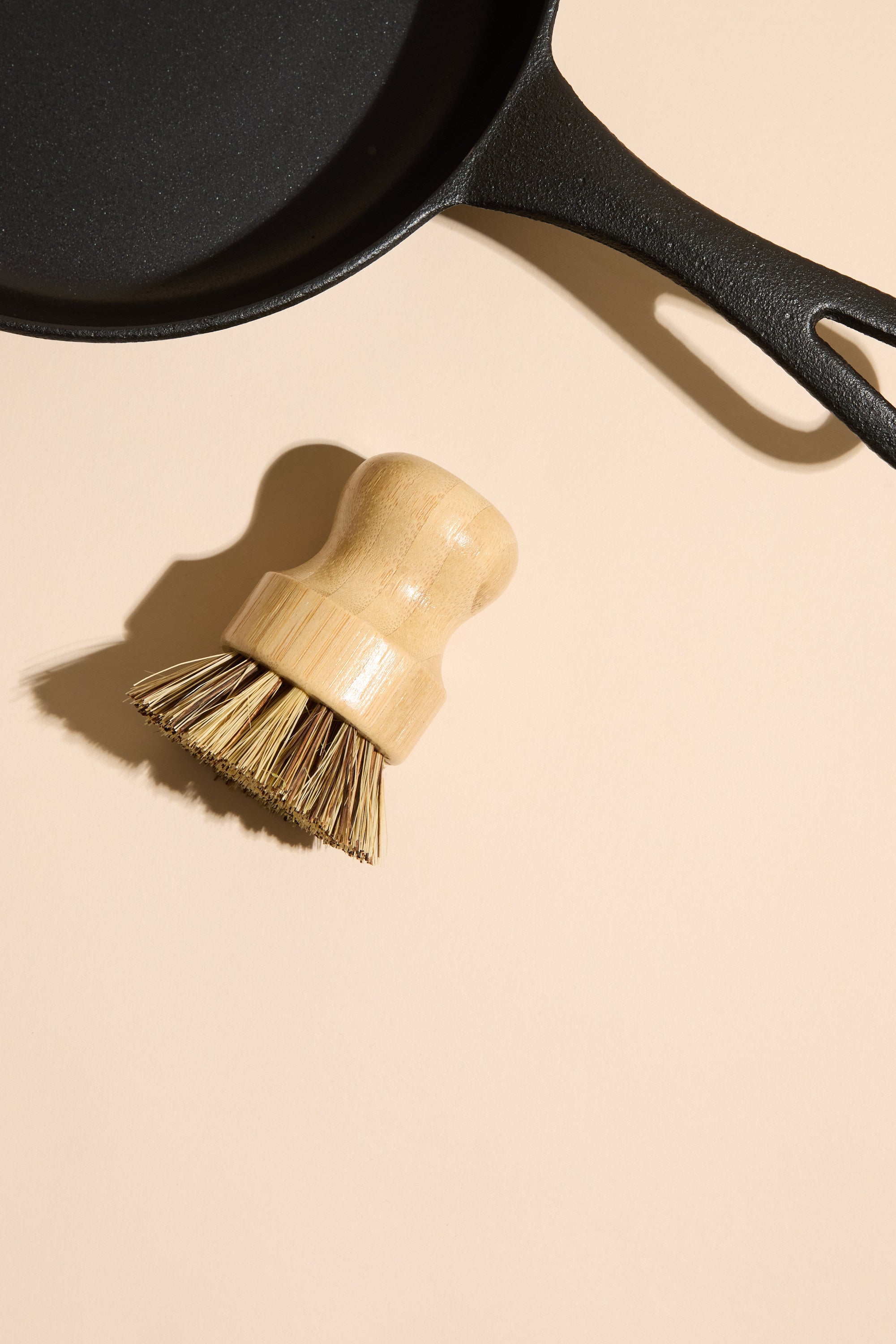 The Ironclad Lil' Legacy Pan by Other and a wooden scrub brush with bristles rest on a light beige surface, their shadows cast to the left.