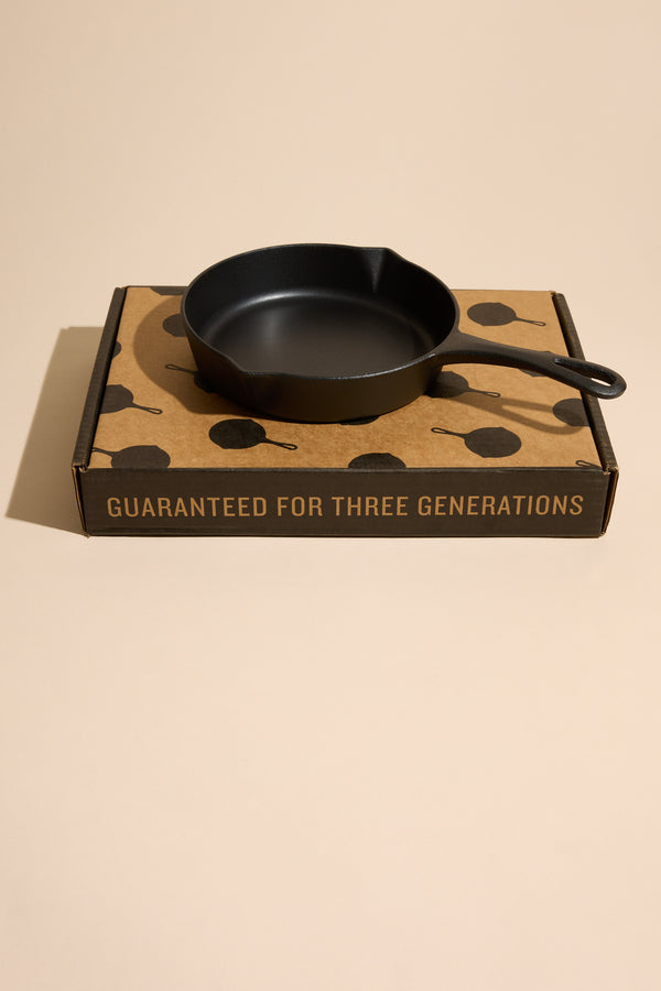 An Ironclad Lil' Legacy Pan by Other rests atop a brown box featuring black skillet illustrations and the phrase "GUARANTEED FOR THREE GENERATIONS" printed on its side.