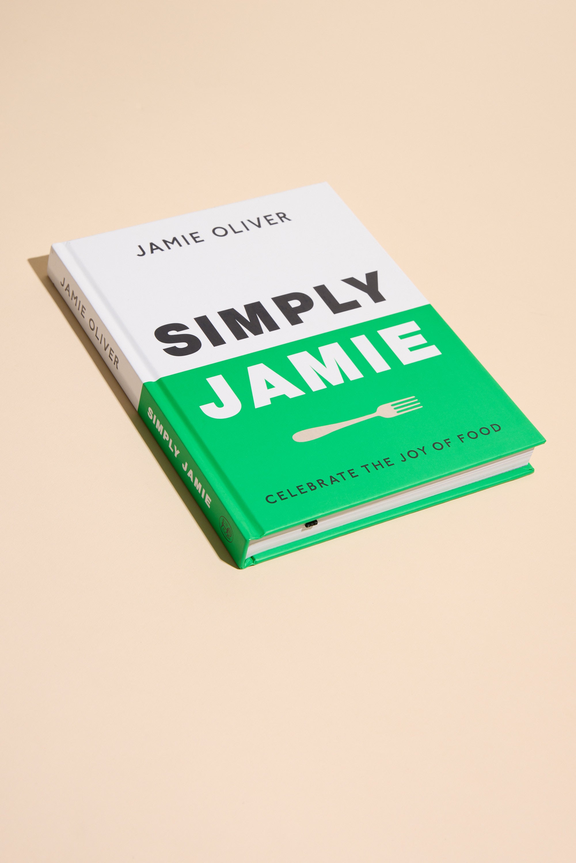 A hardcover cookbook, "Simply Jamie" by Other, with a white and green cover and the subtitle "Celebrate the Joy of Food," rests on a beige surface. It offers easy recipes for tasty midweek meals in bold black and white text.