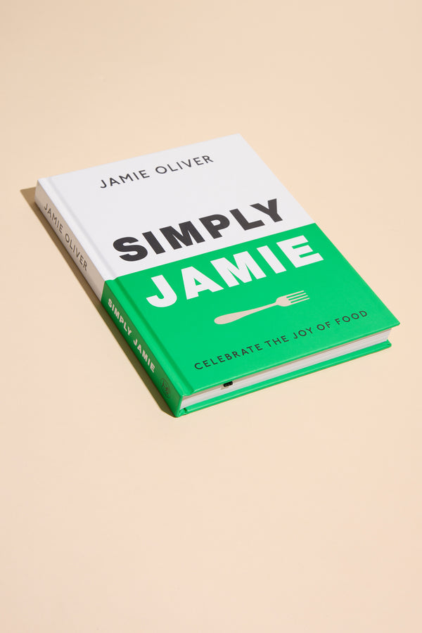 A hardcover cookbook, "Simply Jamie" by Other, with a white and green cover and the subtitle "Celebrate the Joy of Food," rests on a beige surface. It offers easy recipes for tasty midweek meals in bold black and white text.