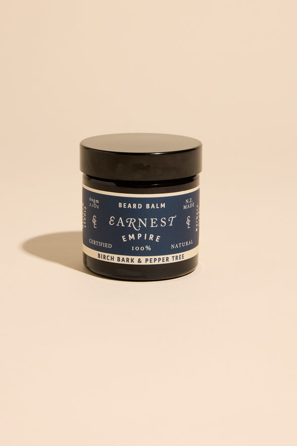 A black-lidded jar of Earnest Empire Beard Balm by Other rests on a beige backdrop. Its label features the Birch Bark & Pepper Tree scent and 100% natural ingredients, detailed in dark blue and white text.