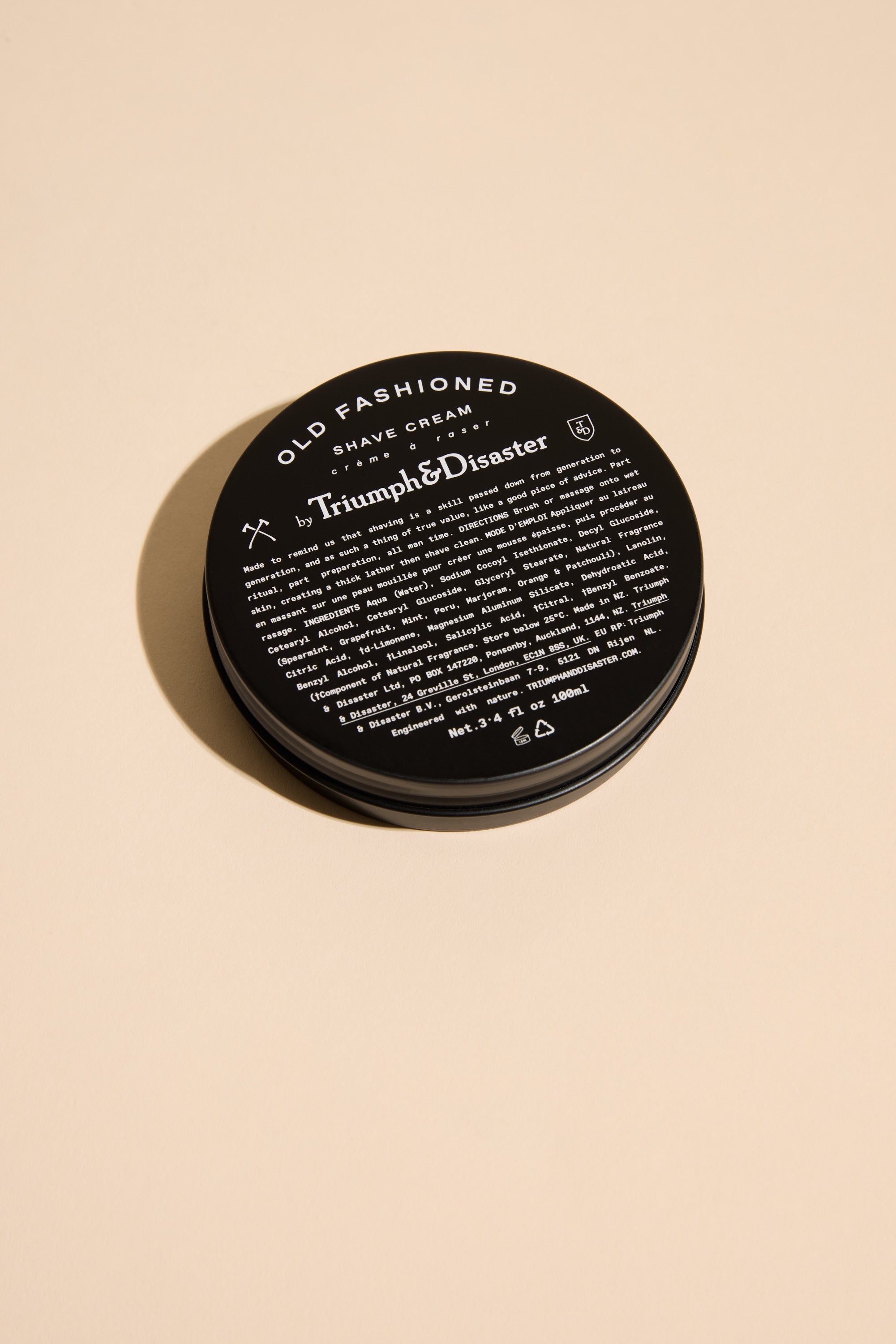 A black round jar of Triumph & Disaster Old Fashioned Shave Cream rests on a beige surface, featuring white text with product details on the lid.