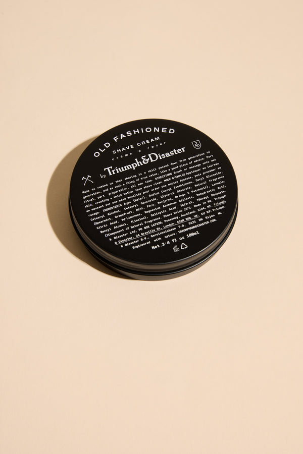 A black round jar of Triumph & Disaster Old Fashioned Shave Cream rests on a beige surface, featuring white text with product details on the lid.